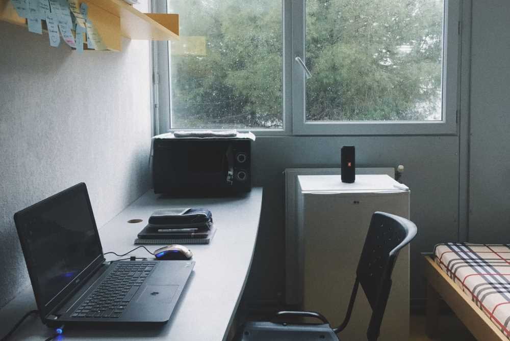 A dorm room containing a desk, microwave, laptop, shelf and bed