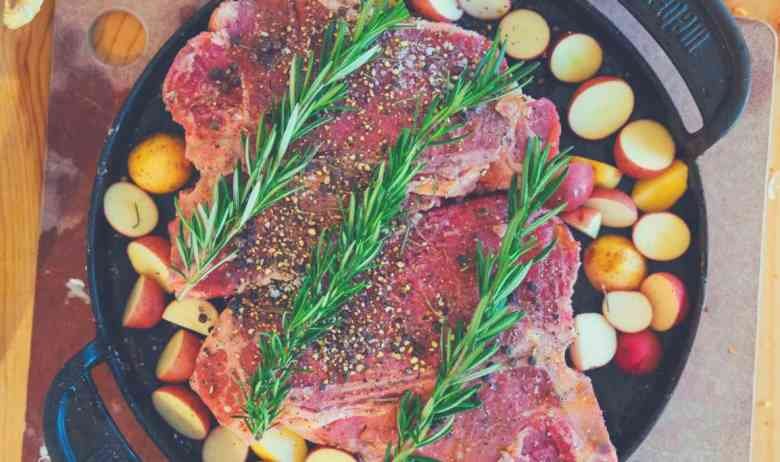 A large cut of meat covered in fresh rosemary and surrounded by vegetables in a skillet