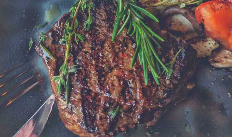 A cooked steak with rosemary on top, surrounded by vegetables