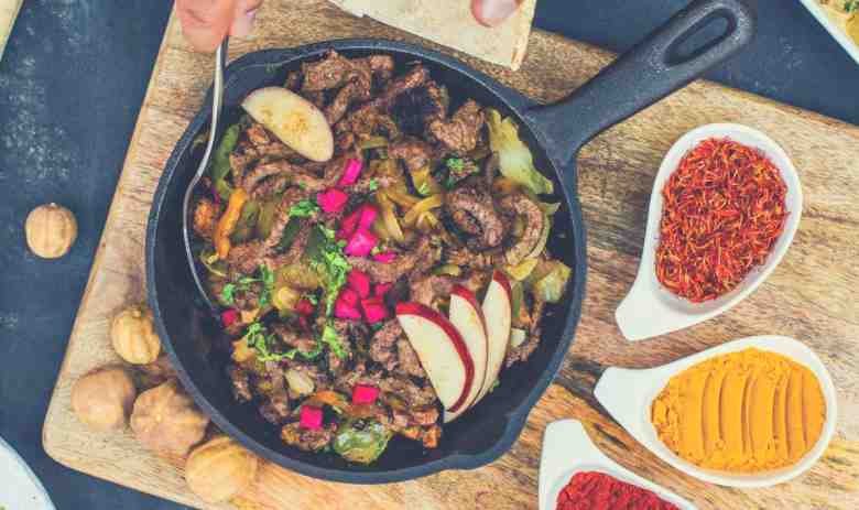 A skillet of meat, fruit and vegetables being served alongside three spices
