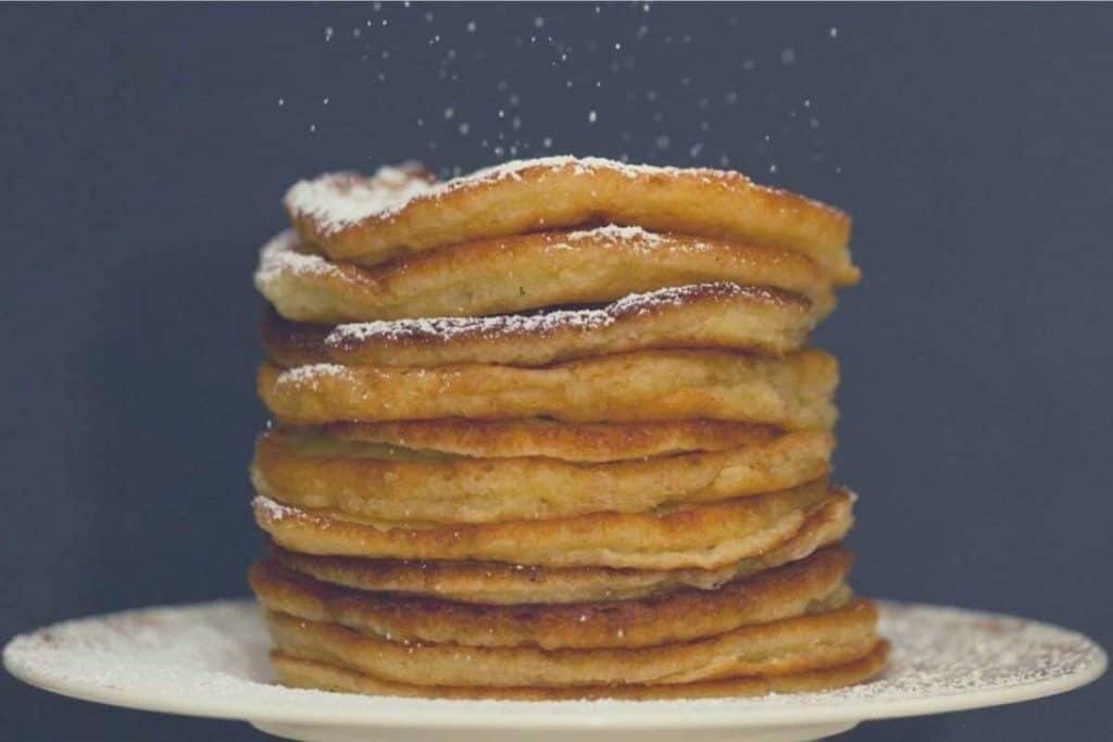 A stack of pancakes sitting on a plate, with icing sugar being sprinkled on top