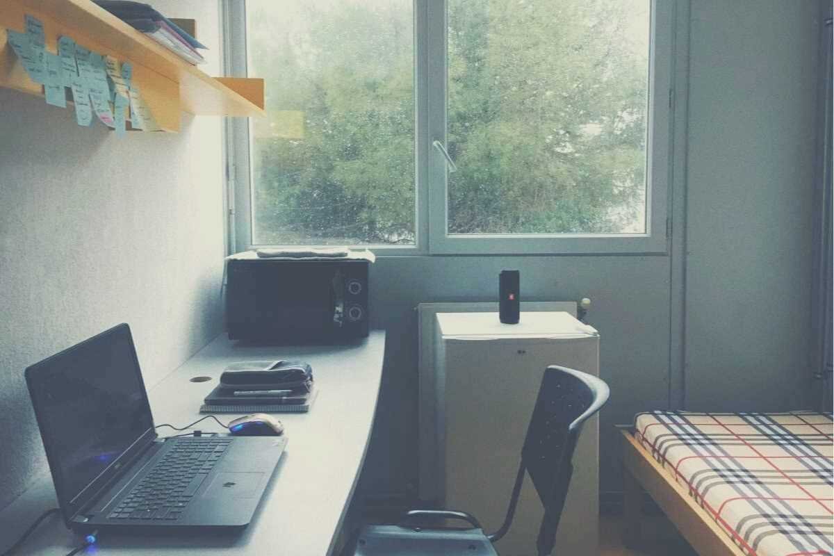 A dorm room containing a window, laptop, desk, shelf, microwave and bed