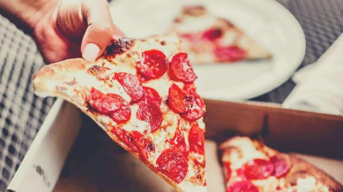 A hand holding a piece of pepperoni pizza, taken from a takeout box