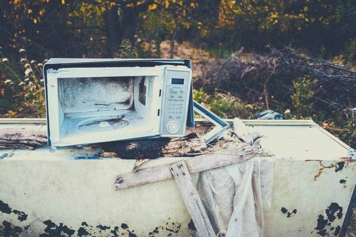 A white broken microwave sitting on a pile of broken furniture in a garden