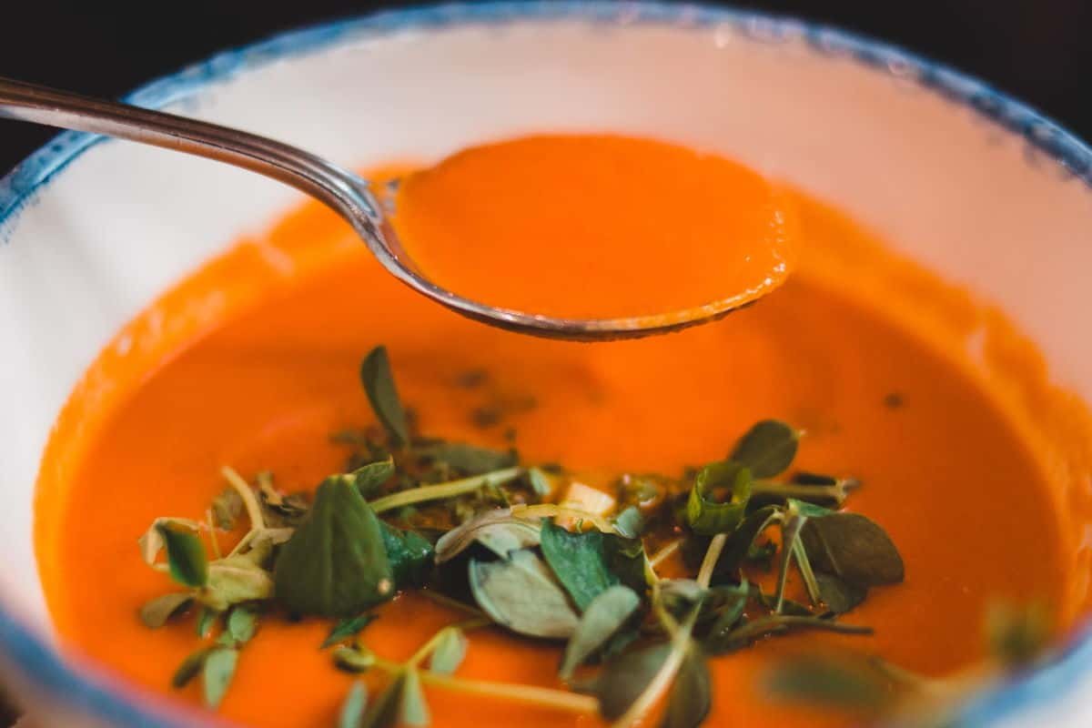 A close up of a bowl of tomato soup and a spoon. There are fresh herbs on top of the soup